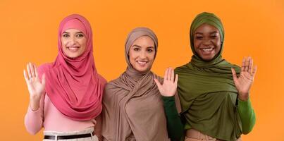 Hello. Three Cheerful Muslim Ladies In Hijab Waving Hands In Greeting Gesture Smiling To Camera Standing Over Yellow Studio Background. Arabic Women Gesturing Hi Or Goodbye. Panorama photo