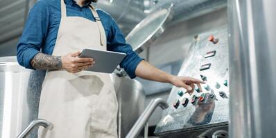 Male worker on beer factory, management and mobile app. Busy male employee or owner in apron with digital tablet controls equipment at brewery and push button, cropped, free space, panorama photo