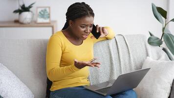 Problem With Internet. Frustrated black lady using laptop and talking on cellphone at home, calling customer support hotline, suffering connection failure while working remotely at home, free space photo