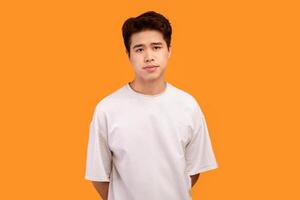 Male Model. Portrait of asian guy wearing blank white t-shirt for design print mockup, standing isolated over orange studio background. Calm casual teenager posing and looking at camera photo