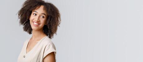 Beautiful model, student, blogger or employee. Happy young african american female with curly hair in casual, turns around and looks at empty space for ad, isolated on light background, close up photo
