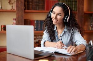 Modern Technology And Elearning. Smiling woman in wireless headset watching webinar online, sitting at desk, using laptop and writing in notebook, taking notes. Education During Quarantine photo