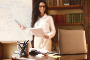 Workspace. Selective focus on laptop on the table, smiling woman in eyeglasses standing at whiteboard in the blurred background. E-learning, remote work, manager giving presentation, online briefing photo