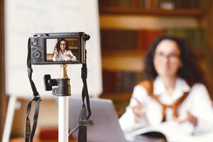 People And Technology Concept. Young woman in glasses sitting at desk, recording broadcast at home, explaining rules, giving remote lecture, talking to camera on tripod for her internet channel photo