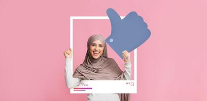A woman wearing a hijab joyfully poses inside a social media frame, giving a thumbs up to celebrate her content engagement. The bright pink background adds to the lively atmosphere. photo