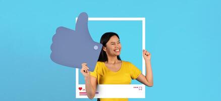 A young woman smiles brightly while posing inside a playful social media frame, holding a large thumbs-up sign. She radiates positivity, embodying the spirit of online content creation. photo