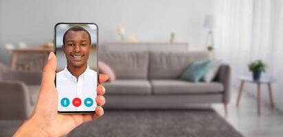 A person holds a smartphone displaying a smiling individual on a call. The setting is a cozy living room, featuring a couch and home decor, perfect for communication. photo