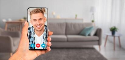 A person holds a smartphone displaying a smiling face during a call. The cozy living room includes a sofa and decorative elements, creating a welcoming atmosphere for communication. photo