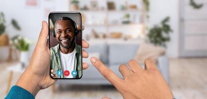 A person is using a smartphone for a call, smiling African America man is on the other end. The background shows a cozy home setting with plants and a couch. photo