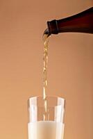 Close-up of golden beer being poured from brown bottle into glass, forming thick foamy head. Captured mid-motion against warm beige background. Beer pouring into glass photo