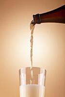 Beer being poured from dark bottle into clear glass with thick foam against warm gradient background photo