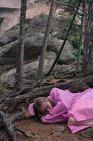 A woman in a flowing pink dress sleeps on the ground under twisted tree roots in a forest with towering rock formations. photo