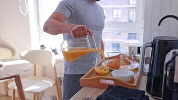 Man pouring orange juice into glasses, preparing breakfast for two with croissants for his wife in a cozy kitchen. video