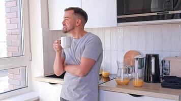 A man drinks coffee in the kitchen at home in the morning. He looks out the window and enjoys a new day. Breakfast at home. Workday or weekend video