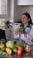 A nutritionist doctor communicates online with patients at a table with fruits and vegetables, explaining the benefits of eating fresh vegetables for the body using green pepper as an example. video