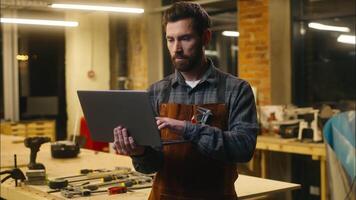 alegre caucasiano cara usando computador portátil dentro oficina carpinteiro artesão madeira mestre artista digitando pc computador feliz construtor masculino surfar Internet homem marceneiro gesticulando polegar acima Boa aprovar aplicativo tecnologia sucesso video