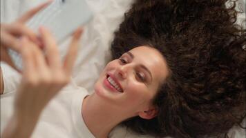 Caucasian woman smiling relaxed girl holding mobile phone on bed wake up morning cheerful happy using smartphone gadget blogger in bedroom weekend texting message browsing cellphone app satisfaction video