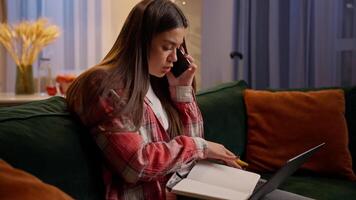 Caucasian multitasking woman studying talk mobile phone writing notes at home night evening online work businesswoman student teen girl using laptop write notebook internet course e-learning on couch video