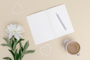 Open notebook, white peony flower, cup of cofffee on table. Creative workspace at home or office for comfort work photo
