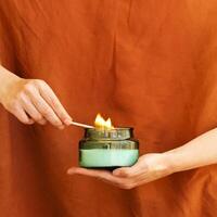 Womans hand uses match to light aromatic candle in recycled glass jar on terracotta color photo