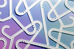 White Plastic hangers as abstract minimal pattern on neon gradient background of purple and blue tint, Clothes rack for organizing clothes in closets or retail stores for hanging of garments, top view photo