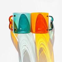 Two Modern Glass Cups with Vibrant sunlight Shadows, Aesthetic Top view of transparent orange and teal color glass mugs for drink. Yellow green reflections, shadow on white, minimal lifestyle photo