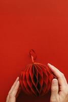 Festive Red Paper Christmas Ornament, hanging red round ball Christmas tree toy with golden shine in female hand on red background, minimal aesthetic life style, festive holiday, top view photo