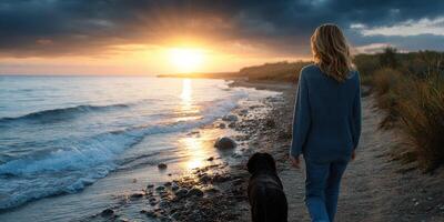 Woman walking her dog along a serene beach during sunset by the water's edge photo
