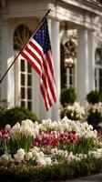 American flag waving in front of a historic building surrounded by vibrant flower beds near sunset photo