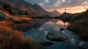 Majestic sunset over serene lake with cross in the foreground amidst towering mountains and lush grass photo
