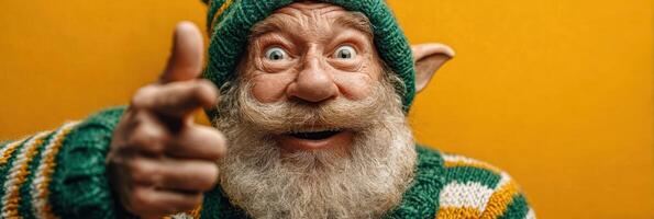Smiling elderly man with a bushy beard and elf costume sitting against a bright yellow background during a festive celebration photo