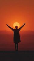 Person stands with arms raised in front of a sunset over distant hills during golden hour photo