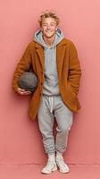 Young man in a cozy coat holding a basketball against a pink wall in urban setting photo