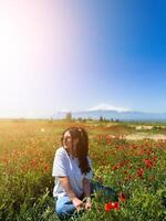 A white girl with curly hair is sitting in a field of red flowers against the background of Mount Ararat in Armenia on a bright sunny day, for an advertisement, for a banner, copy space photo