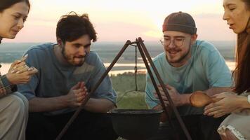 Group of friends, hungry after a long hike, gathered around the campfire. They are watching the cooking of a delicious stew in a pot and are looking forward to it being ready. video