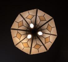 Against a dark background, an octagonal beige lampshade with stained glass windows and three light bulbs hangs from the ceiling, circular pattern. photo