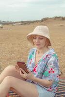 A woman in a sun hat and colorful blouse sits on a beach, holding a phone and looking at the camera under an overcast sky. photo