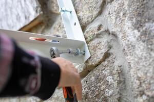 man uses a ratchet wrench with a socket to screw a screw into a metal bracket to install an air conditioner on the stone wall of a rural home photo