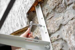 A man uses a ratchet wrench with a socket to screw a screw into a metal bracket to install an air conditioner on the stone wall of a rural home. photo