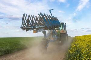 A tractor with a harrow is plowing a field, creating dust clouds under a clear sky during daytime in a rural area photo