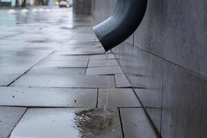 Water drips from a gutter onto rain-soaked pavement, reflecting the gray sky in a quiet city street during a rainy day photo