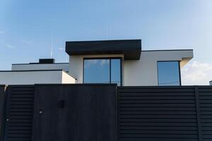 A contemporary house features minimalistic lines and large glass windows, surrounded by a modern fence under a blue sky photo