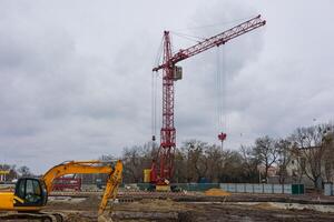 A tall red crane is actively lifting materials over a construction site while heavy machinery is positioned nearby, under a cloudy sky photo