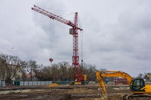 Heavy machinery operates at a construction site featuring a prominent red crane against a gray, cloudy sky, with trees in the background photo