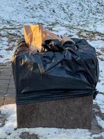 A black trash bag filled with garbage sits atop a stone pedestal near a snowy walkway in a park during late afternoon photo