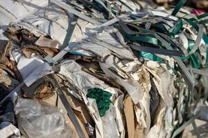 Stacks of shredded cardboard and plastic waste are processed together, creating a tangled mass under bright afternoon sunlight photo