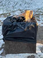 A black trash bag overflowing with waste sits on a concrete base, surrounded by patches of snow in a public area during winter photo