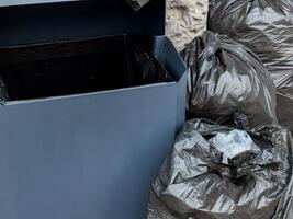 Piles of black garbage bags are placed next to a half-empty trash bin in an urban setting under natural light photo