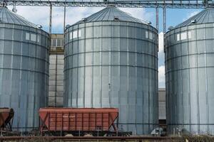Tall metal silos store grain at an agricultural site, with a freight train wagon parked nearby under a blue sky with clouds photo