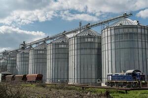 Steel silos used for grain storage stand tall beside a train at an agricultural site under a partly cloudy sky photo